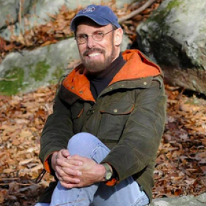 Wendell Minor sitting outside with rocks and fallen leaves in the background.