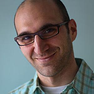 A man wearing glasses in a blue shirt in front of a blue background. 