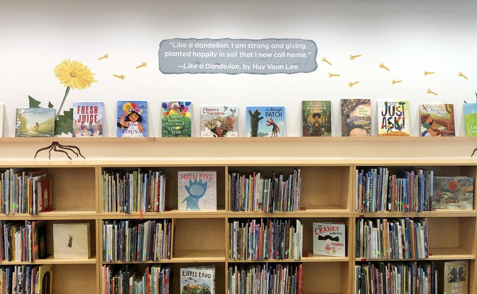 The Rooted Stories shelf in the “Sprouting Stories” Reading Library exhibition. Books displayed on a shelf with dandelions decorating the wall.