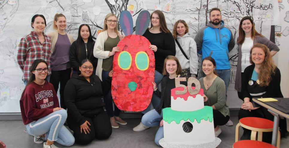 A group of HCC students and educators holding a 50th birthday cake and caterpillar head both made from cardboard and paper.