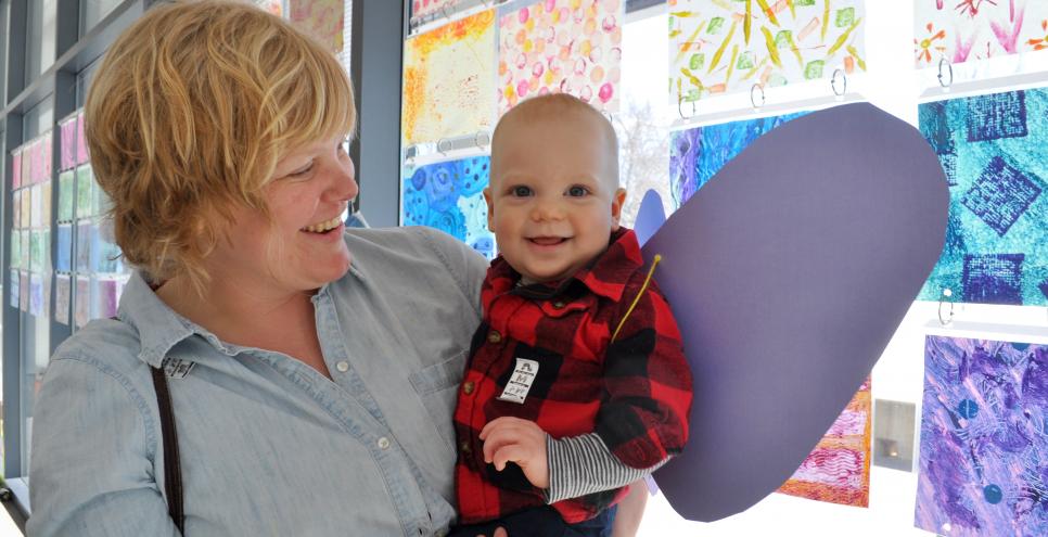 A smiling adult holding a smiling small child wearing purple paper butterfly wings.