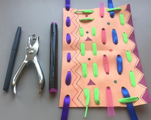 Colorful ribbons woven into orange construction paper. A hole puncher and markers lie next to the weaving.