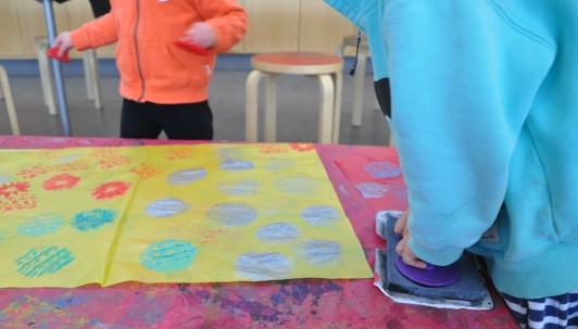 Child stamping with two hands, pressing stamp into silver stamp pad while another child walks around with red stamps in each hand.